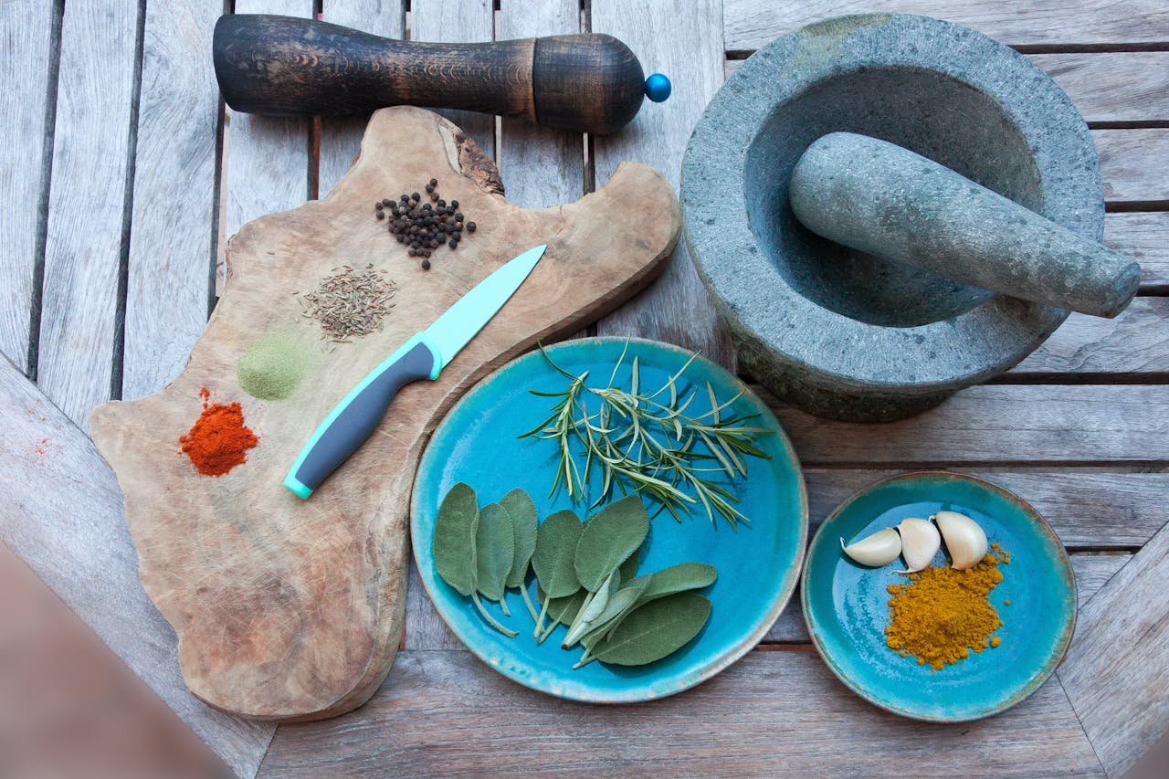 A rustic setup for spice preparation with herbs, spices, garlic, and kitchen tools on wooden table.
